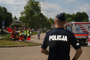 Na zdjęciu widoczny umundurowany policjant. W tle po prawej stronie amulanse, a po lewej ratownicy i strażacy wiozący poszkodowaną osobę. Widoczny również mężczyzna, robiący zdjęcia.
