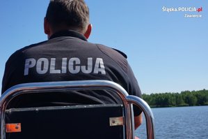 Na zdjęciu widoczny siedzący tyłem umundurowany policjant. W tle widoczny zbiornik wodny.