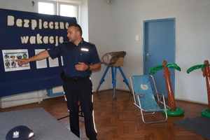 Na zdjęciu widoczny umundurowany policjant prowadzący prelekcję, który wskazuje na jedna kartkę z grafikami, która przypięta jest do tablicy, na której widnieje napis Bezpieczne wakacje oraz przypięte są jeszcze trzy kartki  z tekstem. Po prawej stronie widoczne dwie dmuchane palmy i leżak