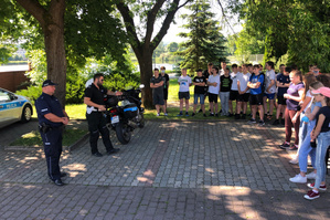 Na zdjęciu widoczni dwaj umundurowani policjanci, podczas prelekcji dla uczniów szkoły podstawowej. Po lewej stronie widać radiowóz oraz policyjny motocykl, po środku i po prawej stronie stoją uczniowie.