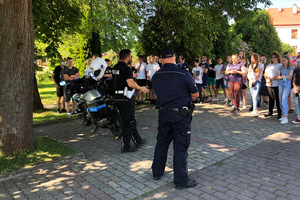 Na zdjęciu widoczni dwaj umundurowani policjanci podczas prelekcji dla uczniów szkoły podstawowej. Widoczni także uczniowie i policyjny motocykl.