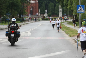 Na zdjęciu widoczny umundurowany policjant jadący policyjnym motocyklem. Po jezdni maszerują uczestnicy marszu nordic walking.