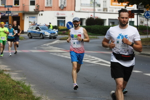 Na zdjęciu widoczni biegnący zawodnicy. W tle widać radiowóz i umundurowanego policjanta, zabezpieczającego bieg.