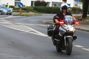 Na zdjęciu widoczny umundurowany policjant jadący policyjnym motocyklem. W tle widoczny radiowóz i policjant.