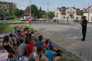 Na zdjęciu widoczny umundurowany policjant, prowadzący spotkanie z siedzącymi na przeciw niego dziećmi. W tle widoczne kolorowe balony.