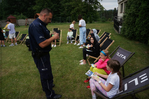 Na zdjęciu widoczny umundurowany policjant prowadzący spotkanie profilaktyczne i rozdaje dzieciom elementy odblaskowe. Dzieci siedzą na leżakach