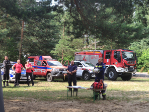 Na zdjęciu widać policjanta i strażaka którzy prowadzą zajęcia profilaktyczne. Z lewej strony stoją strażacy a obok nich stoją pojazdy służbowe.