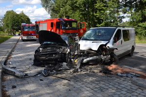 Na zdjęciu widoczne dwa zniszczone wskutek zderzenia samochody. W tle wozy strażackie i strażacy.