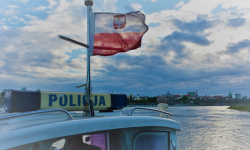 Na zdjęciu widoczna flaga Polski umieszczona na dachu policyjnego statku. Flaga widoczna jest na tle zbiornika wodnego.