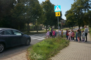 Umundurowany policjant wraz z dziećmi przechodzą przez jezdnię po pasach. Widoczny jest także samochód osobowy, który zatrzymał się przed pasami.