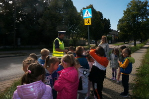 Umundurowany policjant tłumaczy dzieciom oznakowanie przejścia dla pieszych, przy którym stoi wraz z nimi.