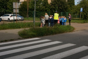 Umundurowany policjant stojąc wraz z dziećmi przy przejściu dla pieszych, tłumaczy im oznakowanie tego miejsca