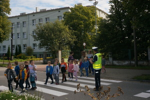 Dzieci przechodzą przez jezdnię po pasach. Policjant stojąc na jezdni obserwuje ich zachowanie