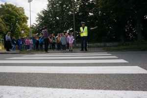 Umundurowany policjant stojąc przed przejściem dla pieszych tłumaczy dzieciom, jak poprawnie przechodzić przez jezdnię