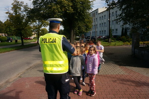 Umundurowany policjant stojąc na chodniku wraz z dziećmi, tłumaczy im, jak zachować się w rejonie przejścia dla pieszych