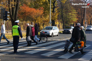 Na zdjęciu widoczny umundurowany policjant kierujący ruchem w rejonie przejścia dla pieszych, przez które przechodzą osoby. W tle widoczne zatrzymane samochody.