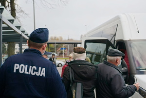 Na zdjęciu widoczny umundurowany policjant wraz z przedstawicielką Sanepidu, idący w kierunku busa. Widoczni również pasażerowie pojazdu, czekający na wejście do auta.