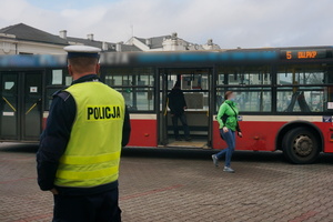 Na zdjęciu widoczny umundurowany policjant obserwujący, czy wysiadający z autobusu pasażerowie mają założone maseczki na usta i nos.