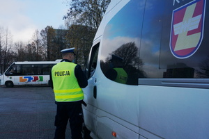 Na zdjęciu umundurowany policjant podczas kontroli, stoi przy drzwiach busa od strony kierowcy. W tle widoczny autobus.