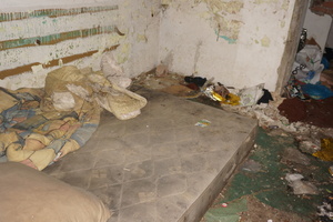 Na zdjęciu widoczny materac, poduszka i kołdra, położone w pustostanie. W tle widoczne śmieci. Ściany pomieszczenia są brudne i odrapane. Panuje ogólny nieład.