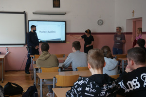 Na zdjęciu widoczny umundurowany policjant oraz pani pedagog, która wskazuje na tablicę z wyświetloną prezentacją. Na tablicy widoczny napis: HANDEL LUDŹMI. Po prawej stroni sali stoi nauczyciel i nauczycielka. Uczniowie siedzą w ławkach.