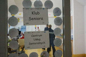 Na zdjęciu widoczne drzwi z przyklejonymi na szybie kartkami, na których widnieją napisy: Klub Seniora. Centrum Usług Senioralnych. W tle widoczni policjanci