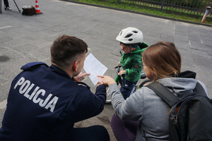 Umundurowany policjant pokazuje dziecku rysunek na kolorowance profilaktycznej. Mama dziecka również wskazuje na malowankę.