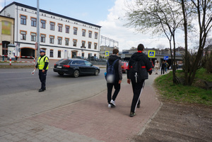 Na zdj zdjęciu widoczny policjant obserwujący ruch uliczny. Po chodniku idą piesi. Osoby przechodzą także przez przejście dla pieszych.