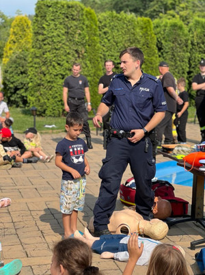 Na zdjęciu widać policjanta u boku którego stoi dziecko i manekiny do pierwszej pomocy, w tle stoją strażacy.