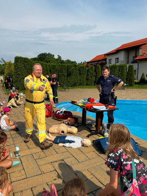 Na zdjęciu widać strażaka i policjanta prowadzącego spotkanie z dziećmi które są na pierwszym planie. W tle widać basen.