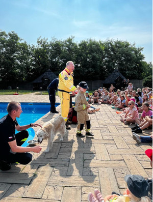 Na zdjęciu widoczny jest basen, strażacy oraz pies służbowy koloru białego. Obok stojąc dzieci.