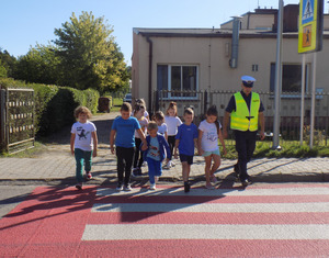 Na zdjęciu widoczny umundurowany policjant ruchu drogowego z grupą dzieci. Oczekują na chodniku w celu przejścia przez przejście dla pieszych na drugą stroną drogi.