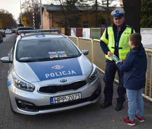 Na zdjęciu widać umundurowanego policjanta, który dziecku wręcza elementy odblaskowe.