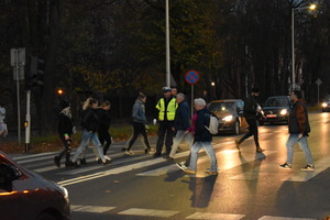 Na zdjęciu widać umundurowanego policjanta który na drodze kieruje ruchem.  Przez przejście dla pieszych przechodzą osoby