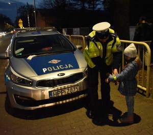 Na zdjęciu widać umundurowanego policjanta, który dziecku wręcza elementy odblaskowe.  W tle widać stojący radiowóz.