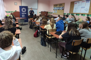 Na kolorowym zdjęciu widoczni uczniowie siedzący w ławkach. Umundurowany policjant prowadzi prelekcję.