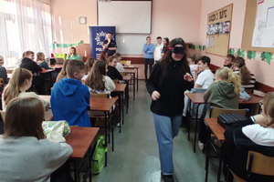 Na kolorowym zdjęciu widoczni uczniowie siedzący w ławkach. Umundurowany policjant prowadzi prelekcję. Obok niego stoi troje uczniów. Uczennica w narkogoglach na głowie przechodzi pomiędzy ławkami.