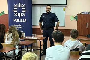 Na kolorowym zdjęciu widoczni uczniowie siedzący w ławkach. Umundurowany policjant prowadzi prelekcję.