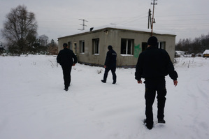 Zdjęcie kolorowe. Policjant i strażnicy miejscy idą po śniegu w kierunku pustostanu.