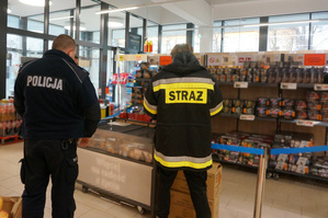 Zdjęcie kolorowe. Policjant ze strażakiem kontrolują stoisko z fajerwerkami.