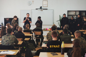 Zdjęcie kolorowe. Widoczny na nim policjant podczas spotkania z młodzieżą. Uczniowie ubrani są w mundury.