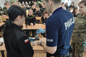 Zdjęcie kolorowe. Widoczny na nim policjant podczas spotkania z młodzieżą. Uczniowie ubrani są w mundury. Policjant oprósza przedmiot trzymany w ręku przez dziewczynę, pędzlem i proszkiem.