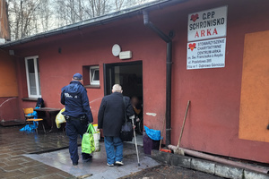 Na kolorowym zdjęciu widoczny jest umundurowany policjant, który niesie torbę z rzeczami idącego obok mężczyzny. Mężczyzna porusza się o kuli ortopedycznej.