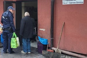 Na kolorowym zdjęciu widoczny jest umundurowany policjant, który niesie torbę z rzeczami idącego obok mężczyzny. Mężczyzna porusza się o kuli ortopedycznej.