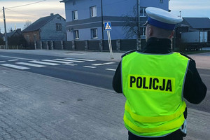 Na kolorowym zdjęciu widoczny umundurowany policjant obserwujący rejon przejścia dla pieszych.