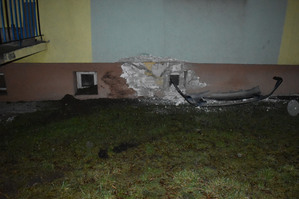 Na kolorowym zdjęciu widoczny częściowo blok mieszkalny z uszkodzoną elewacją. Pod murem widoczne elementy rozbitego pojazdu.