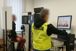 Na kolorowym zdjęciu widoczny policjant w kamizelce odblaskowej z napisem POLICJA, który robi zdjęcia zatrzymanemu mężczyźnie. Zatrzymany mężczyzna siedzi na specjalnym krześle, przeznaczonym do robienia zdjęć przez techników kryminalistycznych.