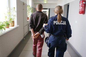 Na kolorowym zdjęciu widoczna policjantka, prowadząca korytarzem zatrzymanego mężczyznę. Mężczyzna na rękach trzymanych z tyłu ma założone kajdanki.
