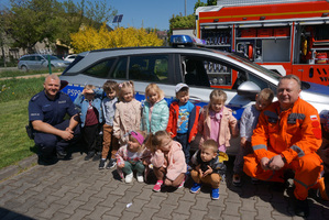 Kolorowe zdjęcie grupowe: policjant wraz z dziećmi i strażakiem. W tle radiowóz.