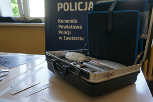 Widoczna teczka technika kryminalistycznego, w tle baner Komendy Powiatowej Policji w Zawierciu.